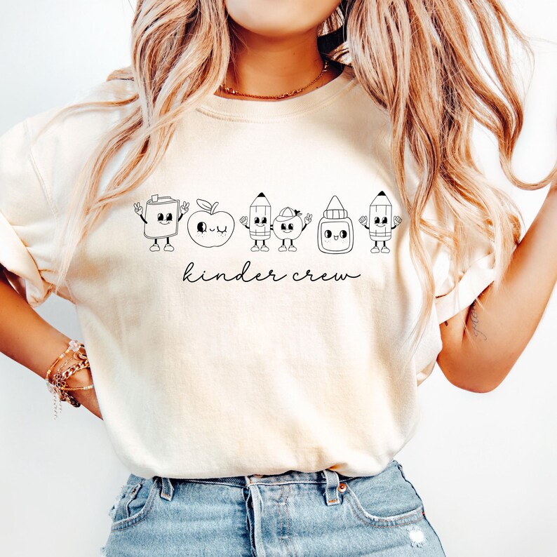 May include: Cream-colored t-shirt featuring a black and white illustration of school supplies with faces, including a glue bottle, apple, and pencils. The words "kinder crew" are written in a script font below the graphic.