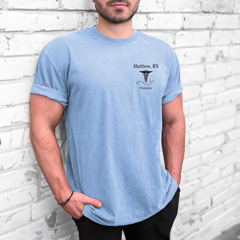 Puede incluir: Una camiseta azul claro con un s&iacute;mbolo de caduceo negro y el texto "Matthew, RN Pediatr&iacute;a".