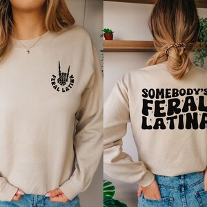Op de afbeelding: Een beige sweatshirt met de tekst "Somebody's Feral Latina" in zwart op de achterkant en een zwart handsymbool met de tekst "Feral Latina" op de voorkant.