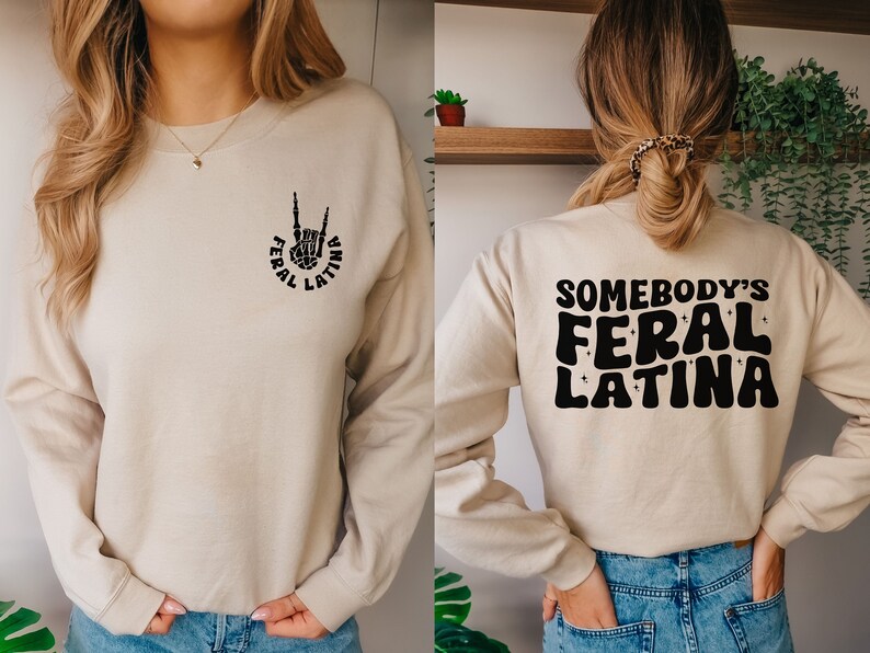 Op de afbeelding: Een beige sweatshirt met de tekst "Somebody's Feral Latina" in zwart op de achterkant en een zwart handsymbool met de tekst "Feral Latina" op de voorkant.
