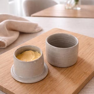 May include: A ceramic butter dish with a lid, filled with yellow butter, sits on a wooden cutting board. Beside it is an empty ceramic container. The butter dish is light gray, and the board has a natural wood grain.