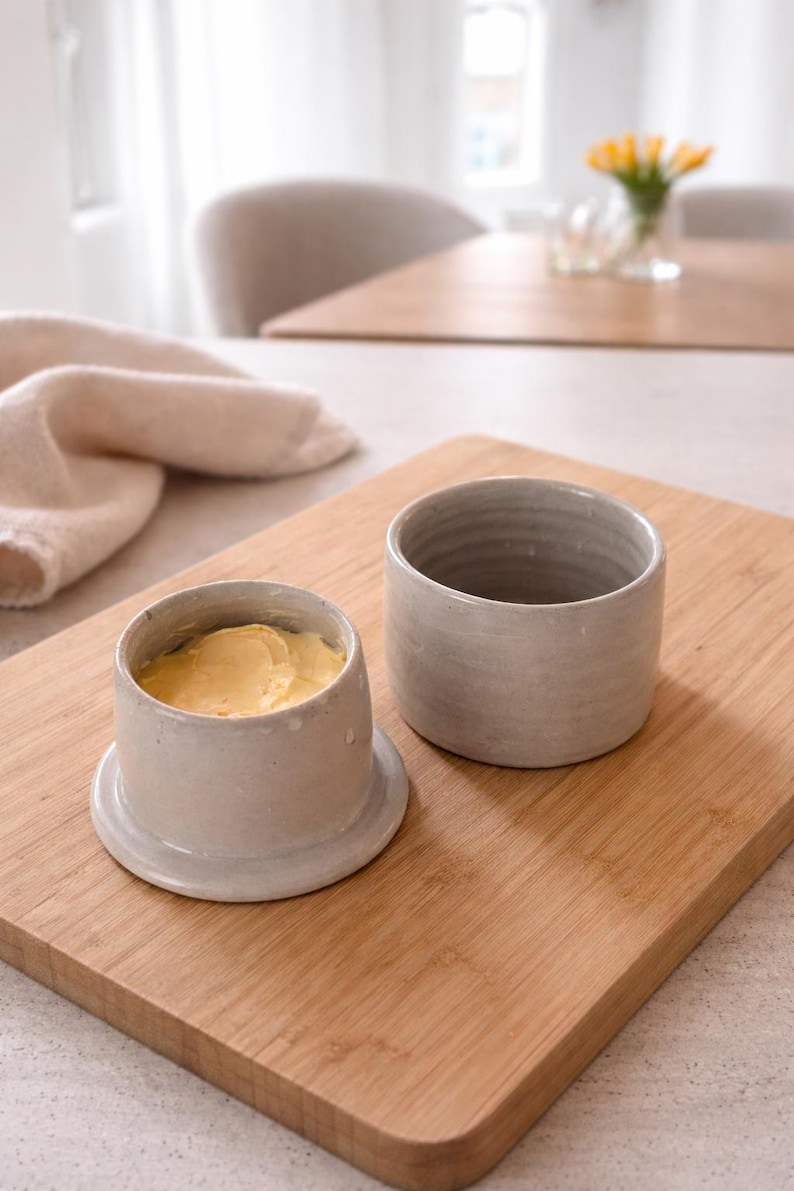 May include: A ceramic butter dish with a lid, filled with yellow butter, sits on a wooden cutting board. Beside it is an empty ceramic container. The butter dish is light gray, and the board has a natural wood grain.
