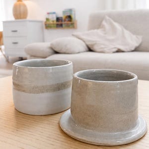 May include: Two ceramic cups in neutral tones sit on a light wood surface. One cup is cylindrical with a wide band of tan. The other cup is a similar shape, but sits on a small, attached base. The background includes a sofa and a lamp.