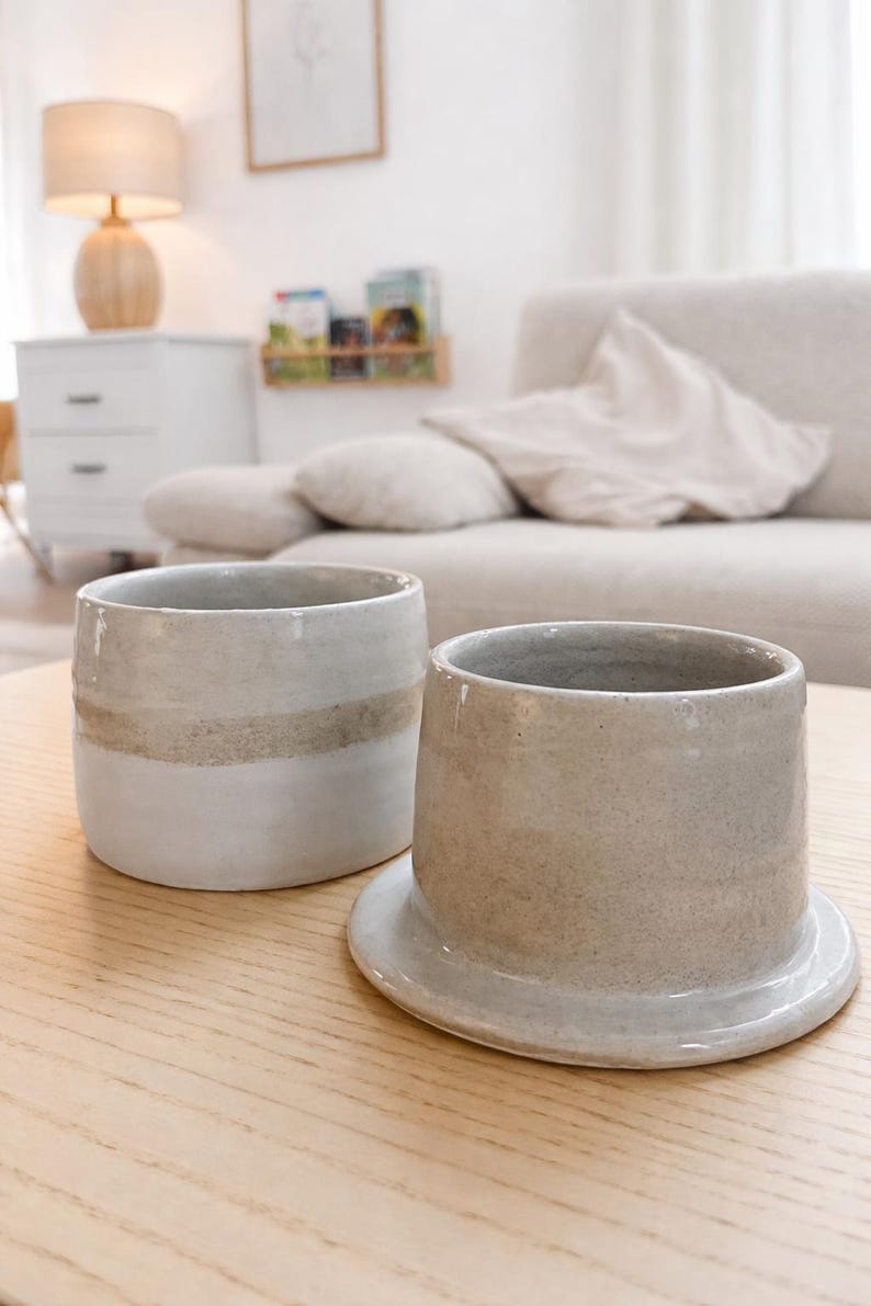 May include: Two ceramic cups in neutral tones sit on a light wood surface. One cup is cylindrical with a wide band of tan. The other cup is a similar shape, but sits on a small, attached base. The background includes a sofa and a lamp.