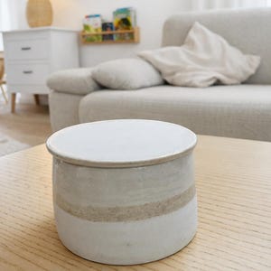 May include: A round, ceramic jar with a lid. The jar is primarily white with a tan stripe around the middle. The jar is sitting on a light-colored wooden table. The background includes a sofa and a lamp.