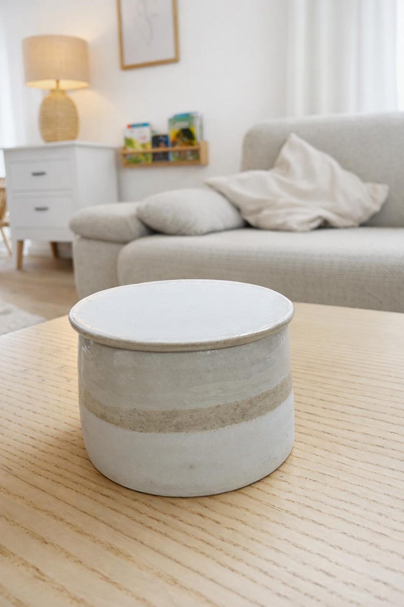 May include: A round, ceramic jar with a lid. The jar is primarily white with a tan stripe around the middle. The jar is sitting on a light-colored wooden table. The background includes a sofa and a lamp.