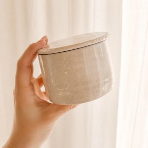 May include: A cylindrical ceramic container with a lid. The container and lid are a light beige color with a textured surface. The container is held in a hand against a soft, neutral background. The container is likely used for storage.