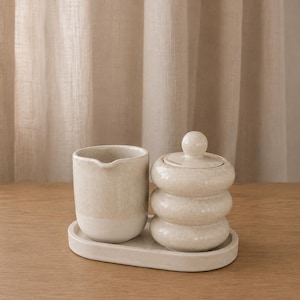 Könnte beinhalten: Ein cremefarbenes Milchkännchen und eine Zuckerdose mit Deckel aus Keramik, beide mit einem gestapelten, abgerundeten Design und einer gesprenkelten Oberfläche. Die beiden Teile stehen zusammen auf einem passenden ovalen Keramiktablett.
