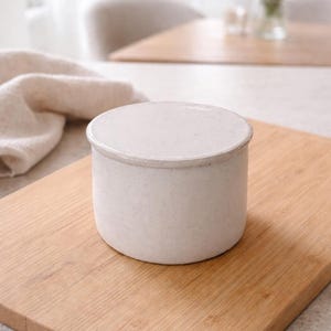 May include: A small, off-white ceramic container with a lid, sitting on a light brown wooden cutting board. The container has a cylindrical shape and a slightly textured surface. The background includes a soft, neutral-toned kitchen setting.