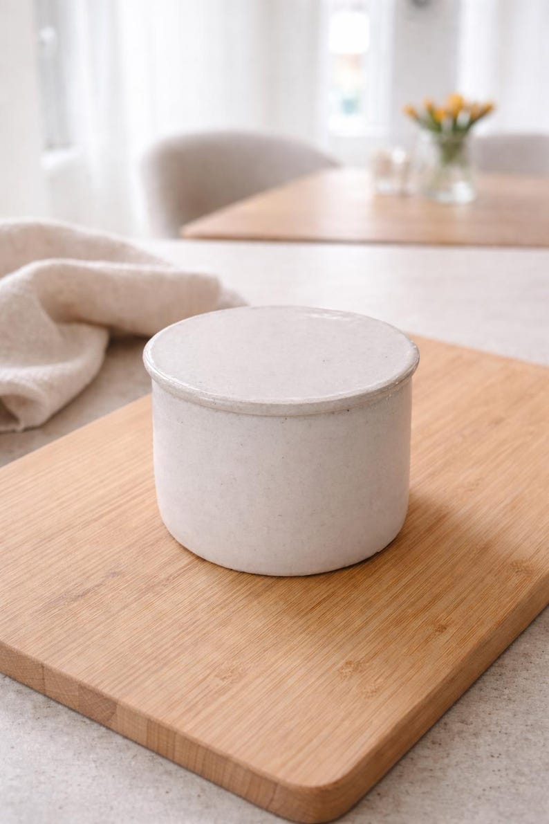 May include: A small, off-white ceramic container with a lid, sitting on a light brown wooden cutting board. The container has a cylindrical shape and a slightly textured surface. The background includes a soft, neutral-toned kitchen setting.