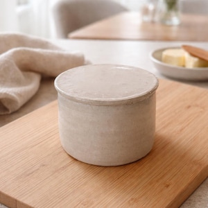 May include: A round, off-white ceramic butter dish with a lid sits on a wooden cutting board. The butter dish is cylindrical and has a slightly textured surface. A folded beige cloth is in the background.