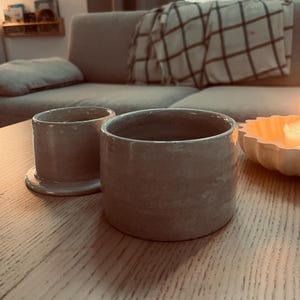 May include: Two handmade, light gray ceramic cups with a textured surface. One cup is cylindrical and sits on a small, matching base. The other cup is also cylindrical, with a slightly wider opening. The cups are on a light-colored wooden table.