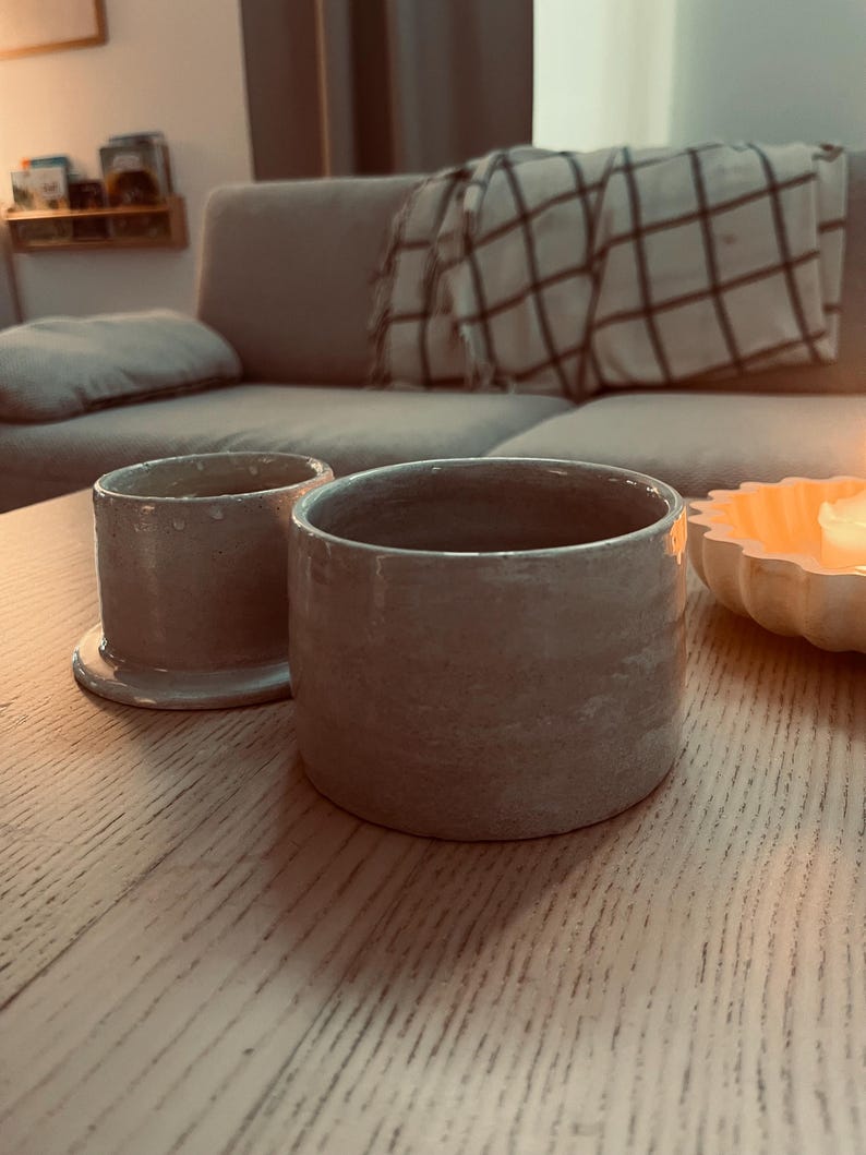 May include: Two handmade, light gray ceramic cups with a textured surface. One cup is cylindrical and sits on a small, matching base. The other cup is also cylindrical, with a slightly wider opening. The cups are on a light-colored wooden table.