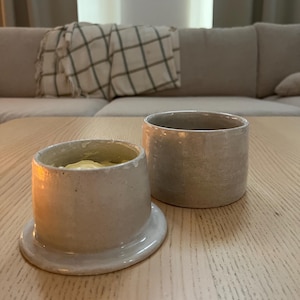 May include: Two ceramic butter dishes in a neutral color palette. One dish is open, revealing butter inside. The other is a simple cylindrical container. Both are on a light wood surface, suggesting a kitchen or dining setting.