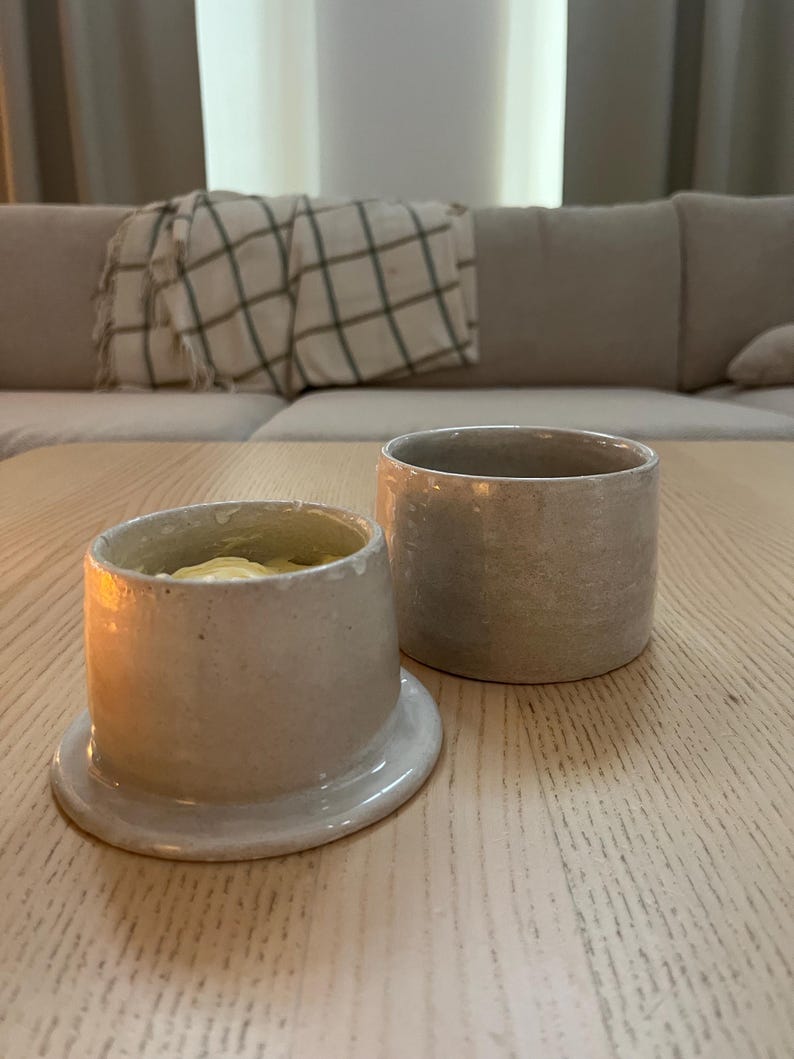 May include: Two ceramic butter dishes in a neutral color palette. One dish is open, revealing butter inside. The other is a simple cylindrical container. Both are on a light wood surface, suggesting a kitchen or dining setting.