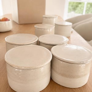 May include: A collection of off-white ceramic jars with lids, arranged on a wooden table. The jars are cylindrical with a slightly textured surface and a contrasting band of color near the base. A cardboard box is open in the background.