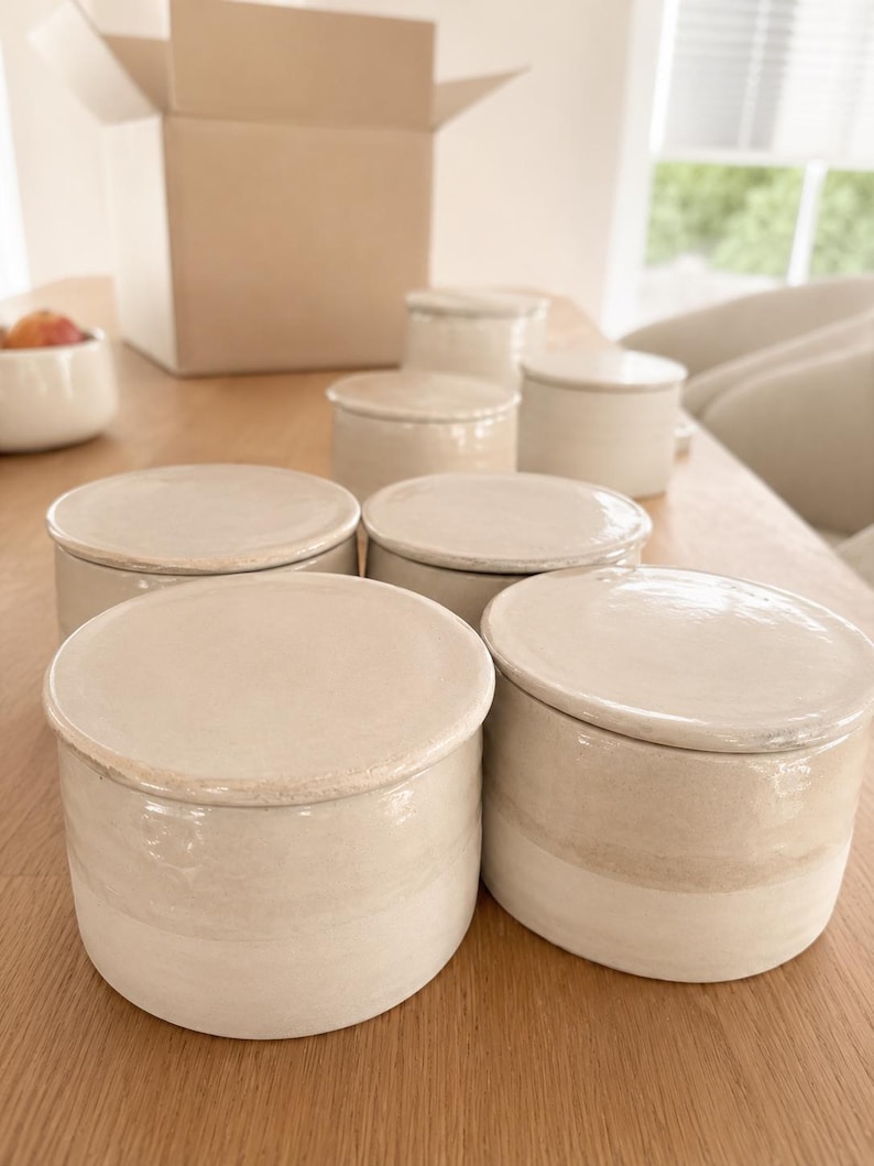 May include: A collection of off-white ceramic jars with lids, arranged on a wooden table. The jars are cylindrical with a slightly textured surface and a contrasting band of color near the base. A cardboard box is open in the background.