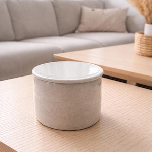 May include: A small, cylindrical ceramic container with a white interior and a light gray exterior. The container sits on a light wood table in a living room setting. The container is a neutral color and could be used for storage or decoration.