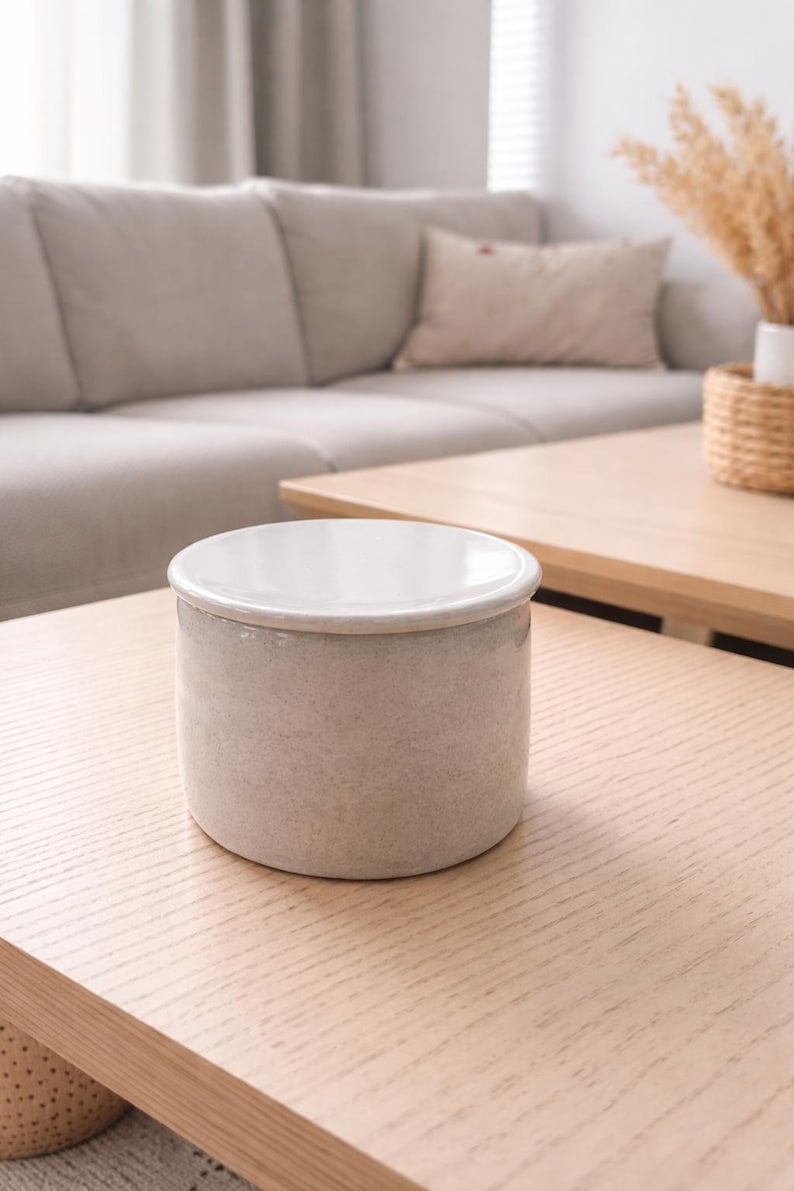 May include: A small, cylindrical ceramic container with a white interior and a light gray exterior. The container sits on a light wood table in a living room setting. The container is a neutral color and could be used for storage or decoration.