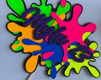 May include: A colorful cake topper with the word "Maya" in purple letters and a neon green, pink, orange, and blue paint splatter design.