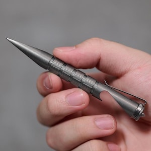 May include: A metallic gray pen with a pointed tip and textured grip. The pen has a built-in bottle opener and a pocket clip. The pen is held in a hand against a gray background.