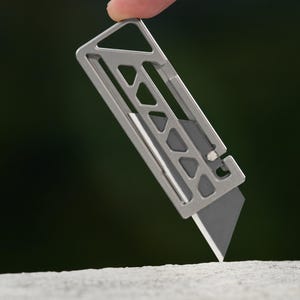 Pode incluir: Um estilete retangular de metal prateado com uma estrutura esqueletizada com recortes triangulares e uma única lâmina afiada exposta na parte inferior. A ferramenta tem um design plano e vazado com um pequeno orifício na parte superior.