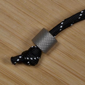 May include: Close-up of a black and white cord with a silver metal bead. The bead has a textured, square pattern. The cord is knotted at the end, and the background is a light brown wooden surface.