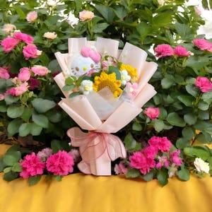 May include: A bouquet of handmade crochet flowers, including a sunflower and white daisies, wrapped in light pink paper. A plush Hello Kitty is nestled among the flowers. The bouquet is tied with a pink ribbon and set against a backdrop of pink flowers and green foliage.