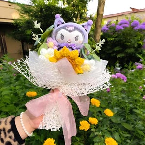 May include: A bouquet featuring a plush character with purple ears and a bow, surrounded by crocheted flowers in yellow, white, and purple. The bouquet is wrapped in white netting and tied with a pink ribbon. The background includes green foliage and yellow flowers.