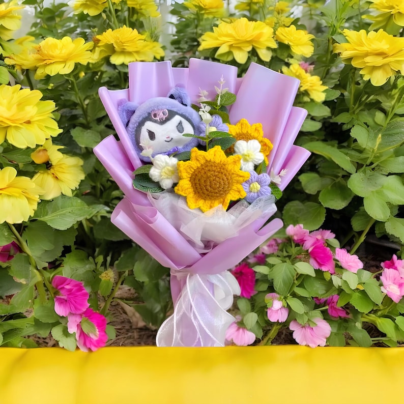 May include: A bouquet wrapped in lavender paper, featuring a plush character, crocheted sunflowers, and other flowers. The bouquet is tied with a white ribbon and set against a backdrop of yellow and pink flowers.