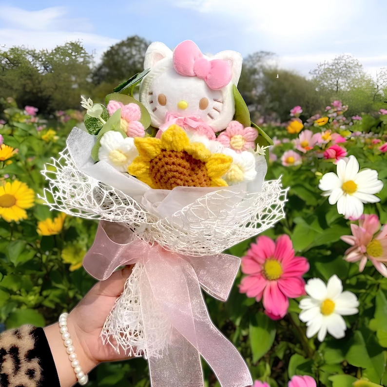 May include: A bouquet featuring a white plush Hello Kitty with a pink bow, surrounded by crocheted flowers and a sunflower. The bouquet is wrapped in white lace and tied with a pink ribbon, set against a backdrop of colorful flowers.