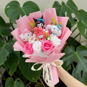 Op de afbeelding: Een boeket verpakt in roze papier met Hello Kitty, Kuromi, My Melody en Cinnamoroll figuren tussen kunstmatige roze en witte rozen en kleine witte bloemetjes, vastgebonden met een wit lint met de tekst Just for you.