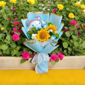 May include: A bouquet featuring a plush character wearing a gray hat, surrounded by crocheted sunflowers and blue flowers. The bouquet is wrapped in light blue paper and tied with a white ribbon, set against a backdrop of colorful flowers.