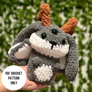 May include: A handmade crochet bunny with antlers. The plush toy is gray with white accents on the face, ears, and belly. It has large, green, glittery eyes and brown antlers. The text "PDF CROCHET PATTERN ONLY" is in a white circle.