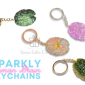Può includere: Quattro portachiavi a forma di cervello umano scintillanti. Due sono verdi, uno è rosa e uno è iridescente. Ogni portachiavi ha un anello dorato e una catena dorata.