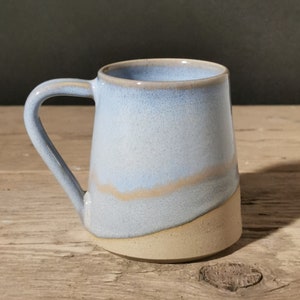 May include: A ceramic mug with a light blue and tan glaze. The mug features a curved handle and a distinctive shape, with a light blue top and a tan base. The mug is on a wooden surface.