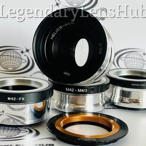 May include: Four silver camera lens adapters with black rings. The adapters are labeled "M42-FX", "M42-NEX", "M42-M4/3", and "HELIOS-44M 2/58".