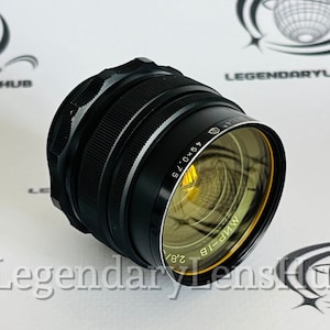 Pode incluir: Uma lente de câmera preta com um anel amarelo ao redor da borda. A lente tem o texto "M.N.P.-18" e "49 x 0,75" impresso nela.