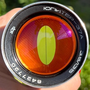 May include: A circular camera lens with a black metal frame and silver trim. The front element features a vibrant orange tint with a vertical yellow oval reflection in the center. Text on the rim reads ЮПИТЕР-37A, 3.5/135, and 8427720.