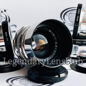 May include: A silver camera lens with a black lens cap and the text "MIR-1 2,8/37" printed on the lens. The lens is surrounded by three other camera lens adapters with the text "M42-FX", "M42-M4/3", and "M42-NEX" printed on them.