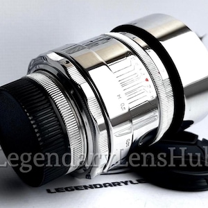 May include: A silver camera lens with a black lens cap. The lens has a silver metal body and a black rubber ring around the front.