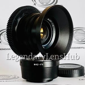 May include: A black camera lens with a gear-like focus ring and a lens hood. The lens is mounted on a black adapter with the text "M42-FX". A lens cap is also present. The text "LegendaryLensHub" is visible.