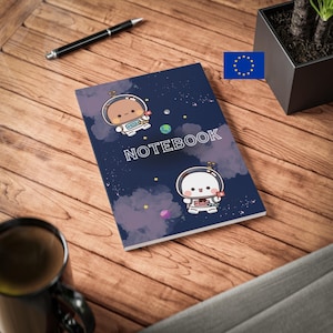 Puede incluir: Una libreta azul con temática espacial con dos astronautas de dibujos animados. La libreta tiene la palabra "NOTEBOOK" impresa en la portada.