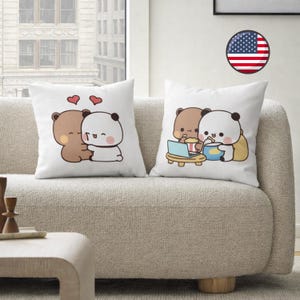 May include: Two white square throw pillows featuring cartoon illustrations of a brown bear and a white panda. One pillow shows the pair hugging with red hearts above them, while the other shows them sitting at a small table with a laptop and snacks.
