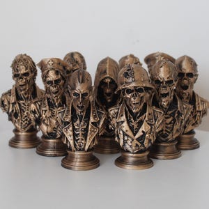 May include: A collection of ten bronze-colored skull busts arranged in a group. Each bust features a unique character design, including figures wearing helmets, hats, and hoods, all with detailed skeletal facial features and textured clothing.