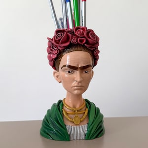 Puede incluir: Un portalápices decorativo con forma de busto de Frida Kahlo. El busto presenta una corona de flores rojas, un chal verde y un collar dorado. Hay bolígrafos de varios colores colocados en el soporte.