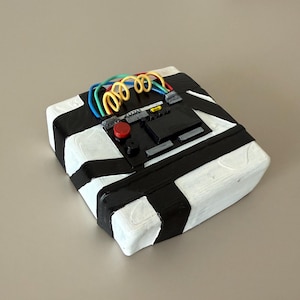 May include: A white box with black tape wrapped around it. The box has a red button and a small circuit board with wires attached. The box is a prop for a film or play.