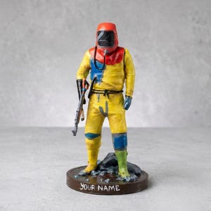 Puede incluir: Una figura de una persona con traje de protección amarillo, capucha roja y visera negra, sosteniendo un rifle. La figura se apoya sobre una base circular con el texto YOUR NAME impreso en blanco en la parte frontal, con detalles en azul y verde.