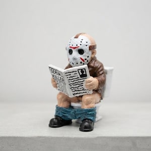Può includere: Una piccola statuetta di un personaggio che indossa una maschera da hockey bianca con segni rossi, una giacca marrone e pantaloni blu abbassati, seduto su un water bianco mentre legge un giornale con il testo SERIAL KILLER in copertina.
