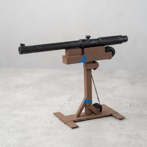 Puede incluir: Un modelo de cañón con un largo tubo negro montado sobre un armón de madera marrón. La estructura incluye un poste de soporte vertical, una base rectangular y pequeñas ruedas negras. Se observan detalles de cinta azul en el marco de madera.